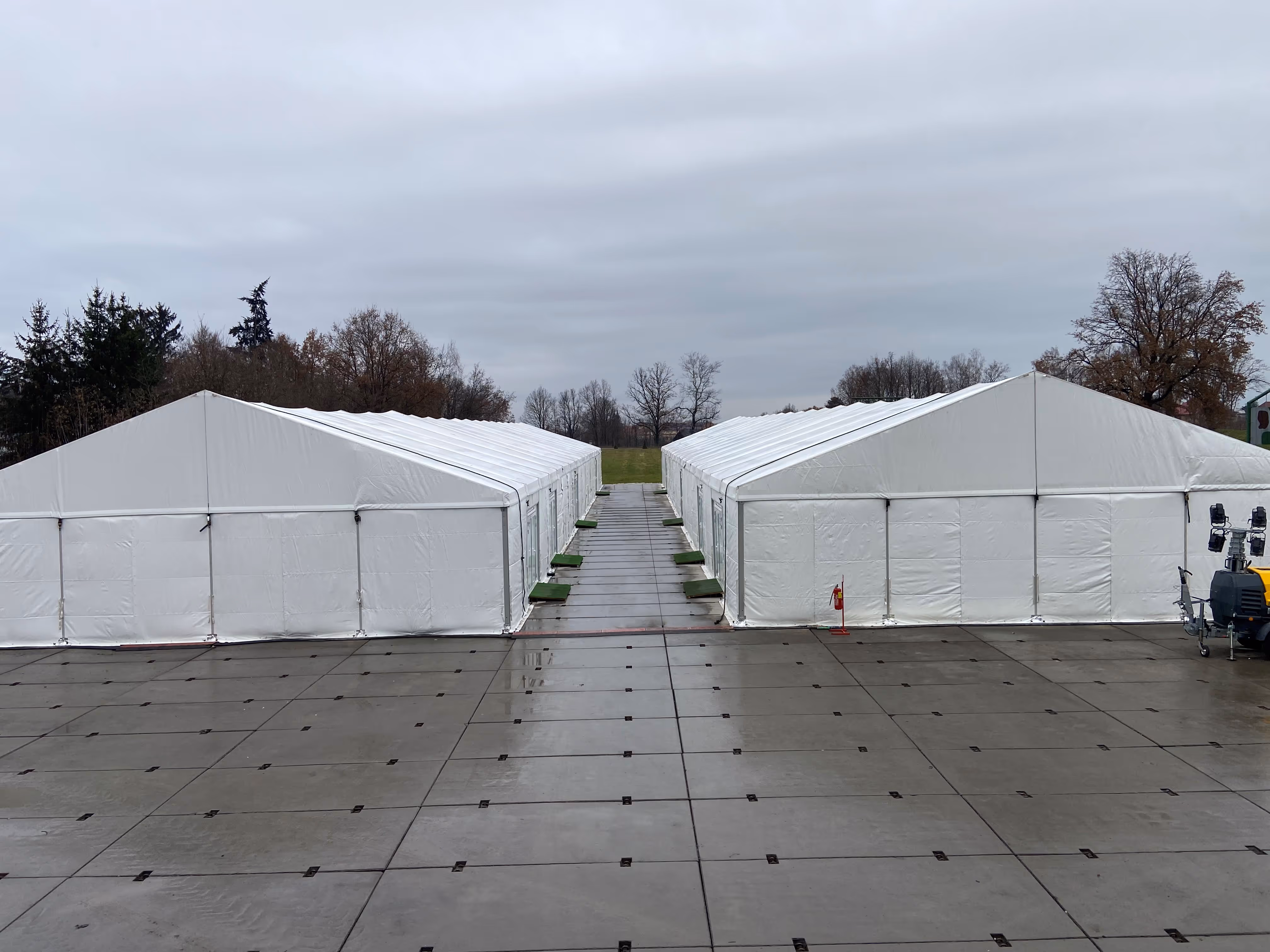 Two large white event tents facing each other on a wet paved surface with overcast sky and trees in the background.
