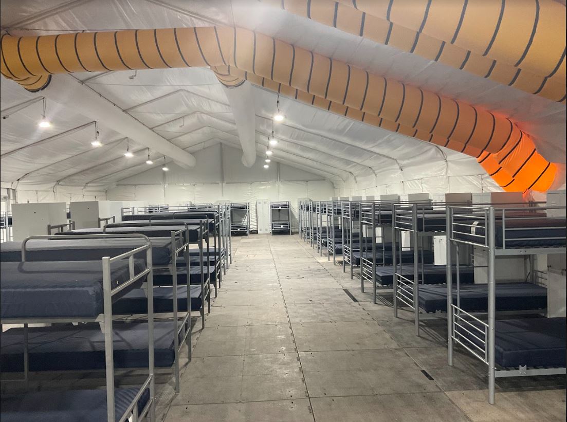 Interior view of a large temporary shelter tent with rows of metal bunk beds and blue mattresses under white fabric ceiling and large orange ventilation ducts.